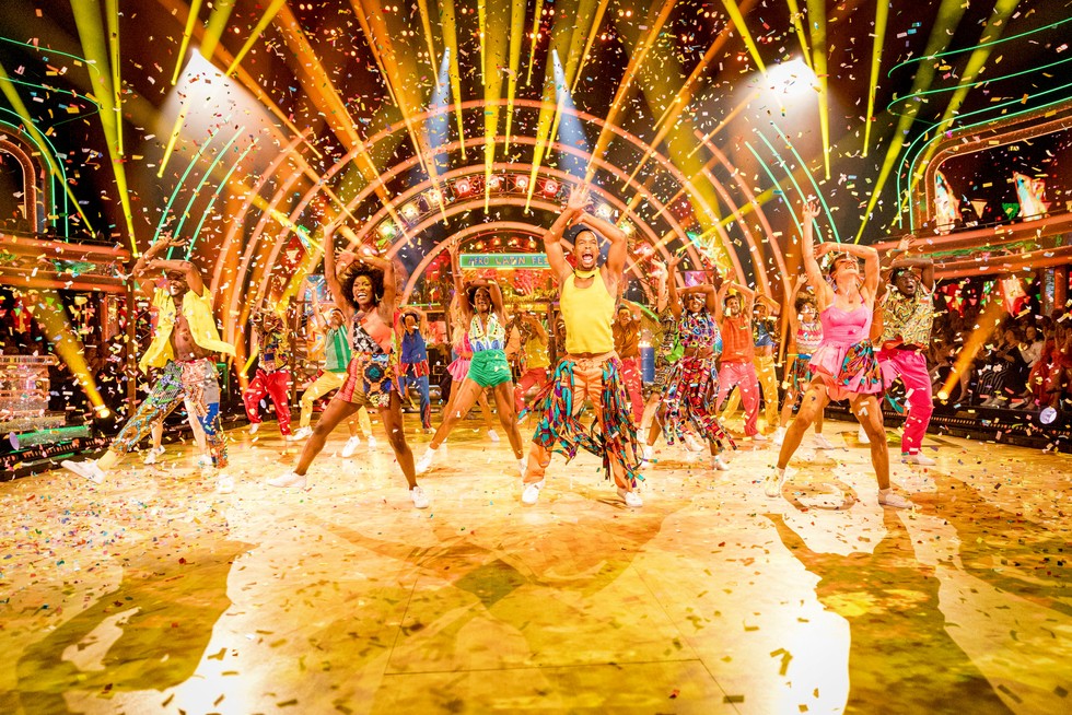 Johannes Radebe leads the professional dancers at the front of the line, wearing a yellow vest and and orange trousers with multi-coloured ribbons hanging from his pockets as he and his fellow pro dancers raise their arms in the air during their opening performance