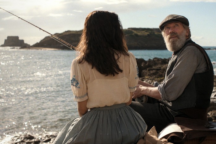 Aria Mia Loberti as Marie-Laure, Hugh Laurie as Etienne LeBlanc in All the Light We Cannot See sitting on rocks overlooking the sea with a fishing rod.