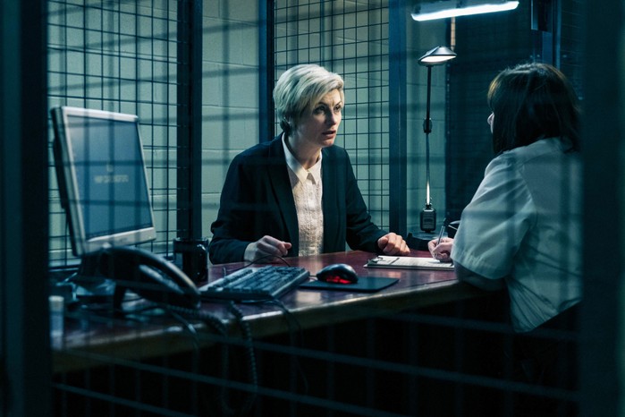 A distressed Orla in prison, standing at a desk, talking to PO Martin